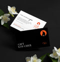 A black gift voucher card with an orange flame logo and white text reading 'GIFT VOUCHER $150' is layered over a white card with product copy and the Candle Supplies logo. The cards are styled on a dark surface with white flowers and green leaves around them.